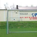 A soccer goal on a green field. In the background, there is a building with the inscription "Sportschule Bitburg."