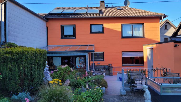 Ein zweigeschossiges, orangefarbenes Haus mit einem gepflegten Garten. Im Vordergrund sind Blumen und Statuen zu sehen.