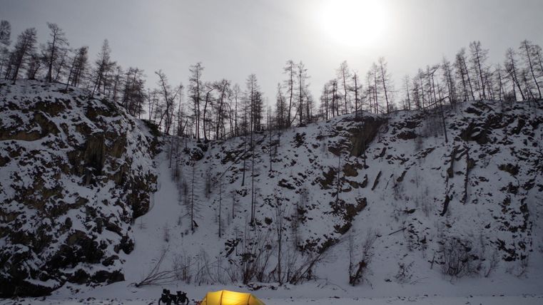 A yellow tent stands in a snowy landscape. In the background, snow-covered mountains and bare trees can be seen, while the sun shines in the sky.