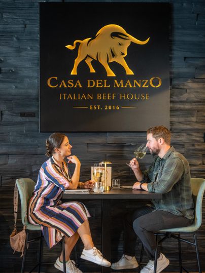 Un couple est assis à une table dans un restaurant nommé "Casa del Manzo". Ils dégustent des boissons et se parlent dans un cadre élégant.