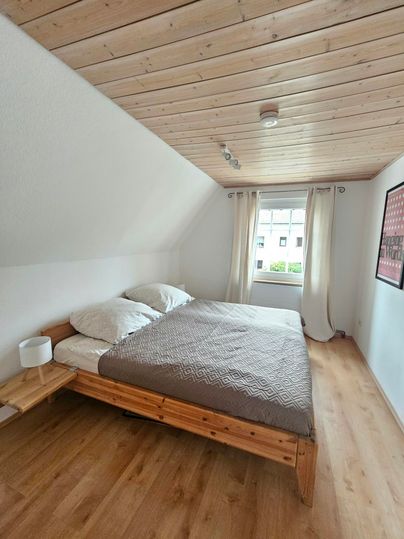 Een gezellige slaapkamer met een houten bed en lichte gordijnen. De kamer heeft een houten vloer en veel natuurlijk licht.