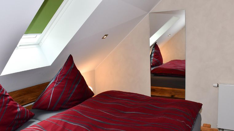 A cozy attic room with a large bed and colorful cushions. A mirror and a skylight provide additional light.