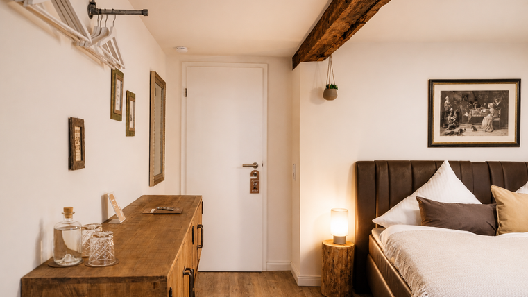 Een gezellige slaapkamer met een groot bed en houten details. Aan de muur hangen schilderijen, en een houten tafel staat naast een fles en glazen.
