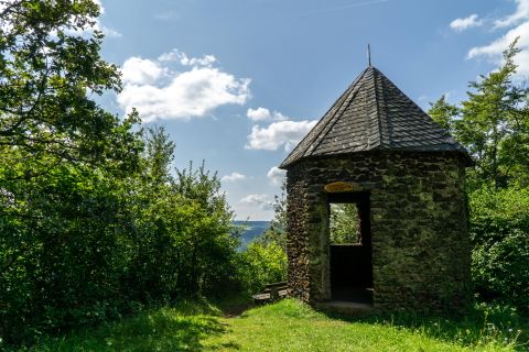 Een stenen paviljoen met een puntdak staat in een groen landschap onder een blauwe hemel.
