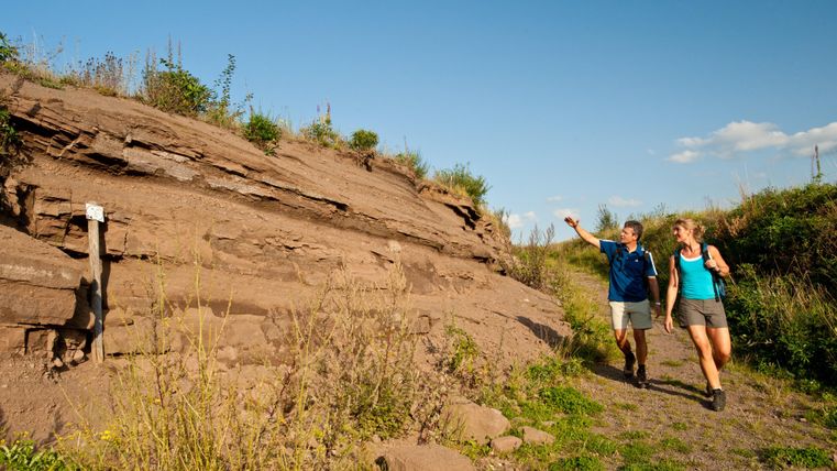Two hikers in summer weather in the Steffeln volcanic garden. One of the two is pointing at the rock face they are walking past.
