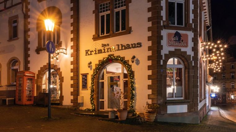 A building decorated for Christmas in Hillesheim at night. String lights and decorations adorn the 'Kriminalhaus' and the 'Café Sherlock'.