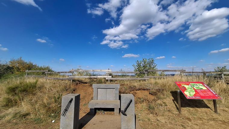 A view of the nature reserve with a concrete bench and an information sign. The sky is clear with some clouds, and the surroundings are green and dry.