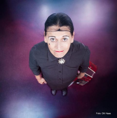 Eine Person in einem schwarzen Kleid steht mit einem Koffer in der Hand. Sie hat eine auffällige Frisur und ein freundliches Gesicht.