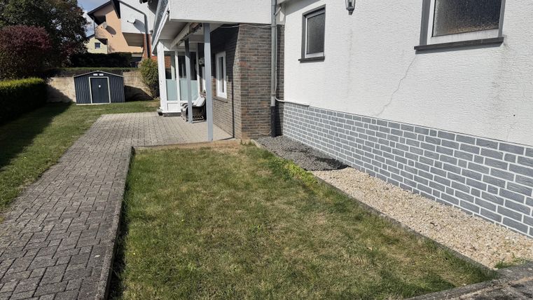 A well-kept outdoor area with a small lawn and paving stones. The wall of the house is light and partially clad with gray stones.