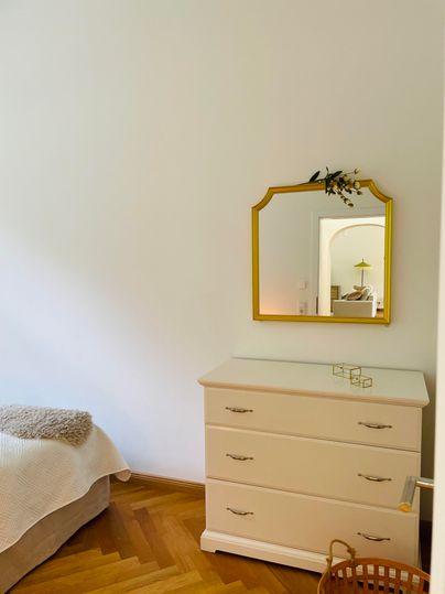 Une chambre lumineuse avec une commode blanche et un miroir encadré en or. Le sol est en bois et il y a quelques éléments décoratifs comme un panier.