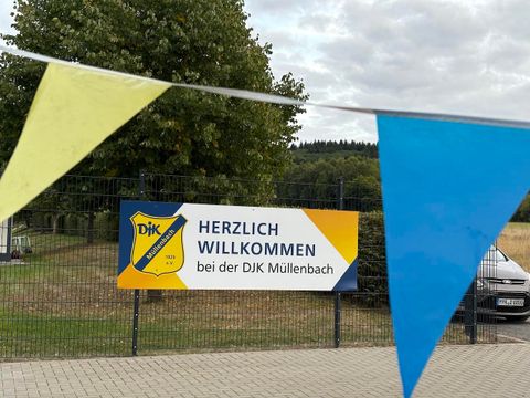 A welcome sign of the DJK Müllenbach, surrounded by colorful flags. The surroundings are green and rural.
