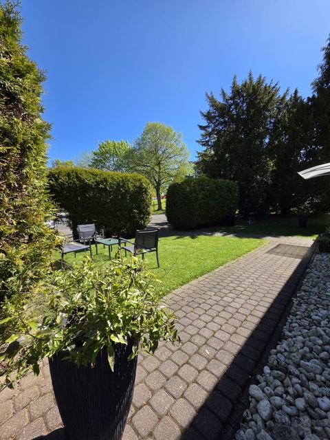Ein schöner Garten mit grünen Büschen und einer hellen Terrasse. Stühle stehen im Freien unter blauem Himmel.