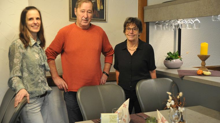 Drei Personen stehen in einem gemütlichen Essbereich. Der Tisch ist dekoriert und es gibt eine warme, einladende Atmosphäre.