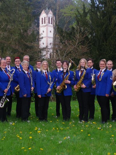 Eine Gruppe von Musikern steht im Freien in eleganten blauen Jacken. Im Hintergrund ist ein Gebäude zu sehen, umgeben von einer grünen Landschaft.