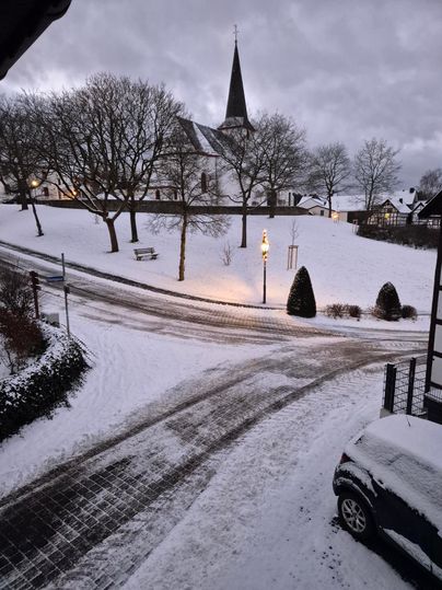 Eine verschneite Landschaft mit einer Kirche im Hintergrund. Die Straßen und Bäume sind mit Schnee bedeckt und es herrscht eine ruhige Atmosphäre.