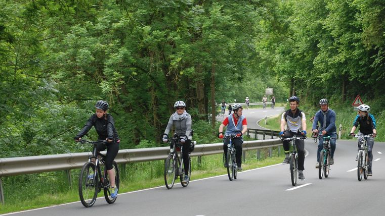A group of cyclists is riding along a country road. The surroundings are green and wooded, creating a peaceful atmosphere.