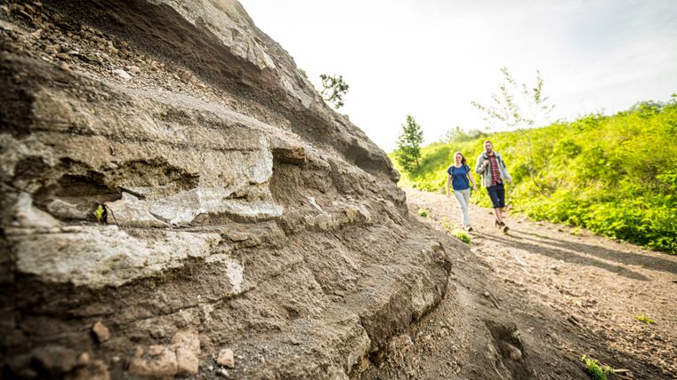 Massive Gesteinsschichten im Vordergrund, an der zwei Wanderer vorbeilaufen.