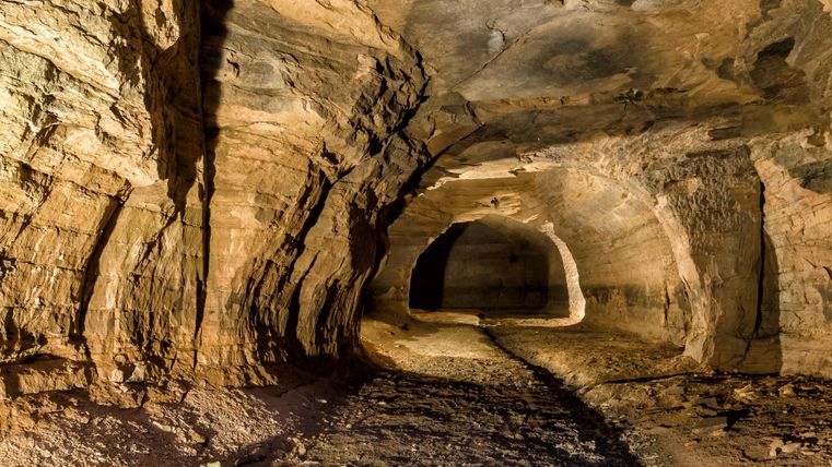 Ein beleuchteter Tunnel mit steinigen Wänden und gewölbter Decke. Der Boden ist uneben und mit Schutt bedeckt. Im Hintergrund ist eine Öffnung sichtbar.