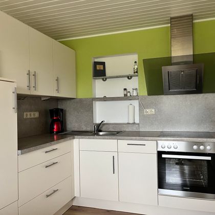 Eine moderne Küche mit weißen Schränken und einer grünen Wand. Sie ist ausgestattet mit einem Herd, einem Kühlschrank und einer Edelstahlabzugshaube.