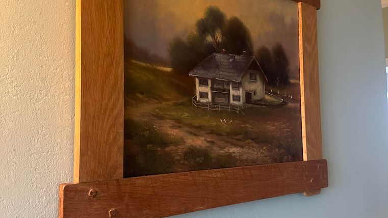 Ein Gemälde eines ländlichen Hauses vor einer ruhigen Landschaft ist an der Wand angebracht. Der Rahmen aus Holz verleiht dem Bild einen rustikalen Charme.