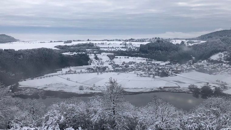 Eine verschneite Landschaft mit einem Fluss und kleinen Dörfern. Der Himmel ist bewölkt und die Natur ist ruhig und still.