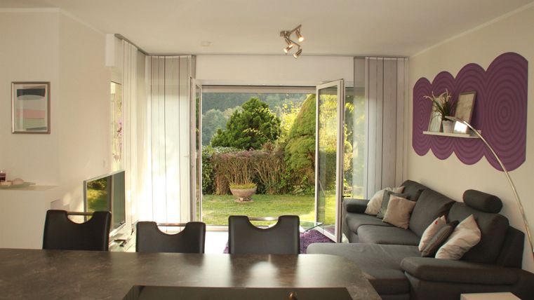 Salon confortable avec un canapé gris et une vue sur le jardin à travers de grandes fenêtres. La pièce a une atmosphère moderne et lumineuse.