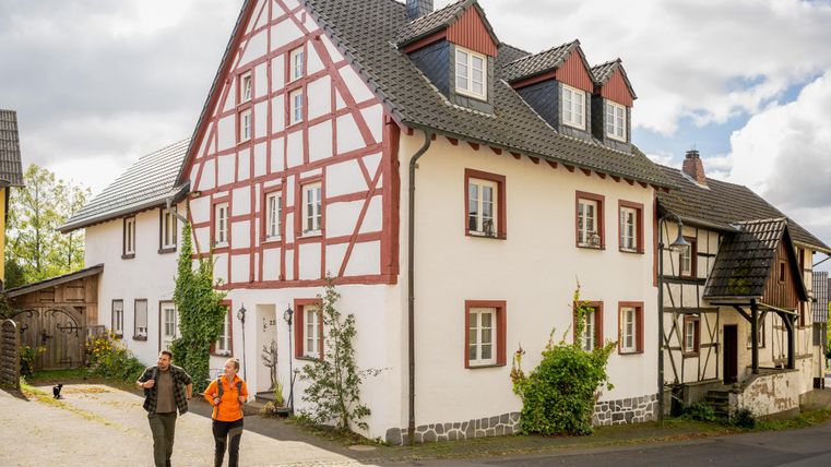 Two people are walking along a traditional half-timbered house. The architecture is typical for a rural area in Germany.