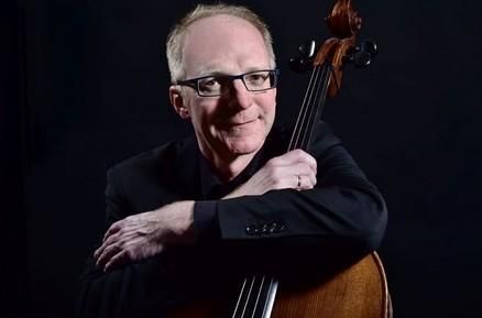Ein Musiker, der im Dunkeln steht und ein Cello umarmt. Er trägt einen schwarzen Anzug und blickt freundlich in die Kamera.