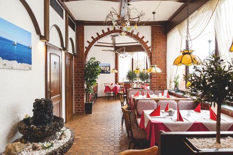 In a room with Mediterranean style, there are several tables with chairs and benches in red, the floor is tiled and there are several plants in the room.