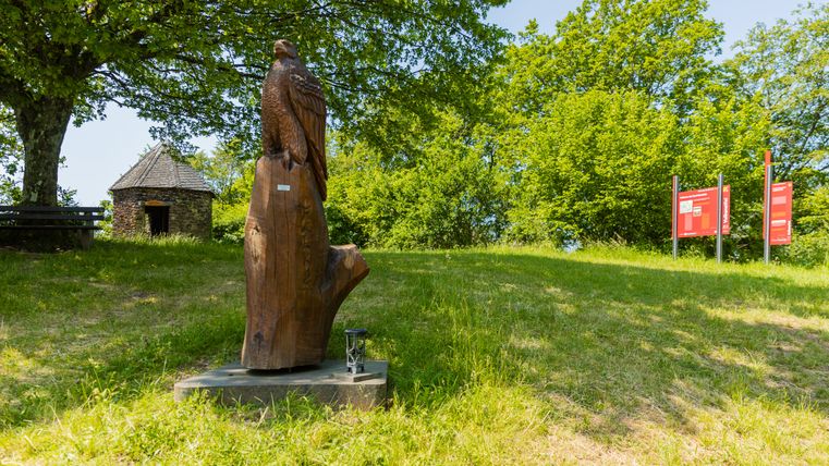 Holzskulptur eines Falken auf einer Wiese mit Bäumen und einem kleinen Steingebäude im Hintergrund.