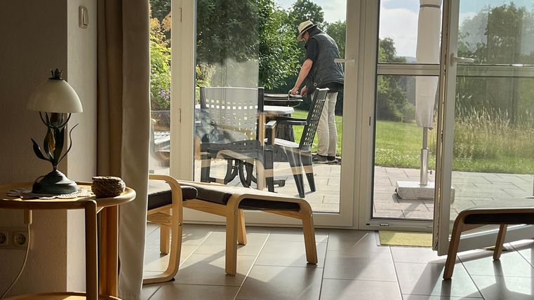 Ein gemütlicher Wohnraum mit großen Fenstern, die auf einen Garten blicken. Im Hintergrund steht eine Person, die an einem Tisch arbeitet.