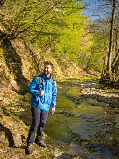A man stands on the bank of a clear stream in a green forest. It is a sunny day and the surroundings feel welcoming and friendly.
