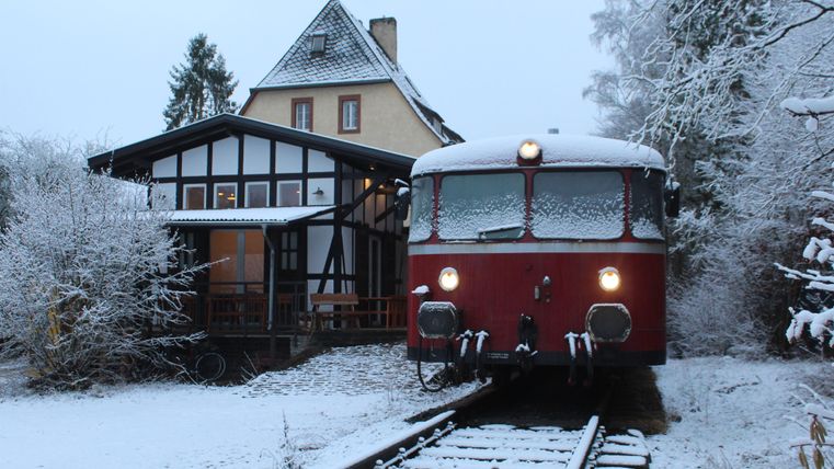 Een rode, met sneeuw bedekte trein staat op de sporen naast een gebouw in een besneeuwd landschap. De scène is rustig en vredig, sneeuw bedekt de grond en de bomen.