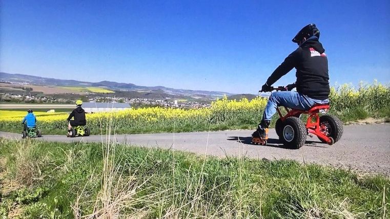 Drei Personen fahren auf kleinen Quads auf einem Weg. Im Hintergrund sind gelbe Rapsfelder und eine hügelige Landschaft zu sehen.