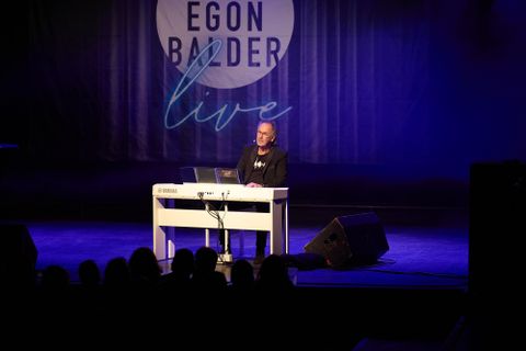 Vier Personen in weißen Outfits sitzen auf dem Boden und posieren vor einer mit Zeichnungen bedeckten Wand. Die Atmosphäre wirkt kreativ und einladend.