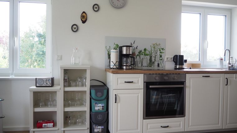 Eine helle, moderne Küche mit Holzregalen und großen Fenstern. Auf der Arbeitsplatte stehen eine Kaffeemaschine und Pflanzen.