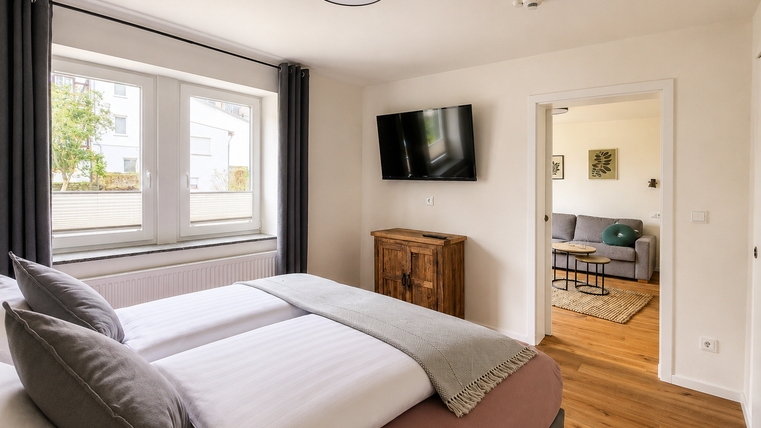 Een moderne slaapkamer met een groot bed en twee ramen. Op de achtergrond is een woonkamer te zien met een bank en televisie.