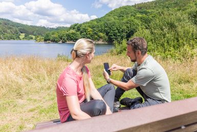 Ein Mann und eine Frau sitzen auf einer Bank am See, mit Blick ins Handy
