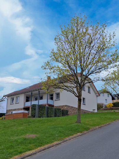 Een modern huis op een helling met een groene grasveld en een boom. De lucht is blauw met hier en daar wat wolken.