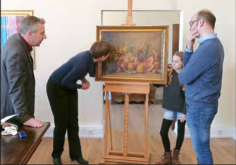 A group of four people is examining a still life-like painting of fruits. They are standing around the easel and showing interest in the artwork.