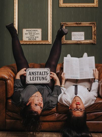 Two women are reading newspapers and a magazine upside down on a leather sofa.