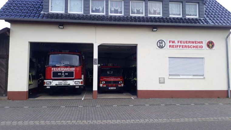 Eine Feuerwache mit zwei Feuerwehrfahrzeugen in den Garagen. Das Gebäude hat ein blaues Dach und ein Schild mit der Aufschrift "FW. FEUERWEHR REIFFFERSCHEID".