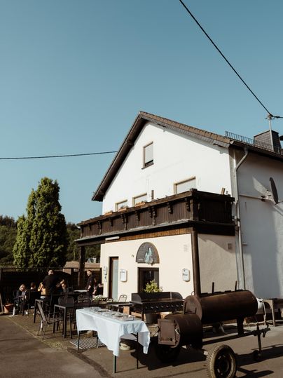 Ein gemütliches Haus mit einem Balkon und einem Gartentisch im Freien. Im Vordergrund steht ein Grill und Menschen genießen die Zeit im Freien.