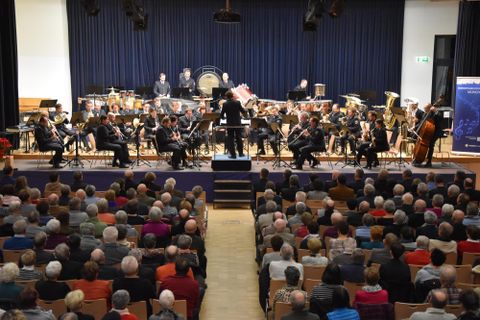 An orchestra is playing on a stage in front of a full audience. The conductor stands in the center and directs the musicians.