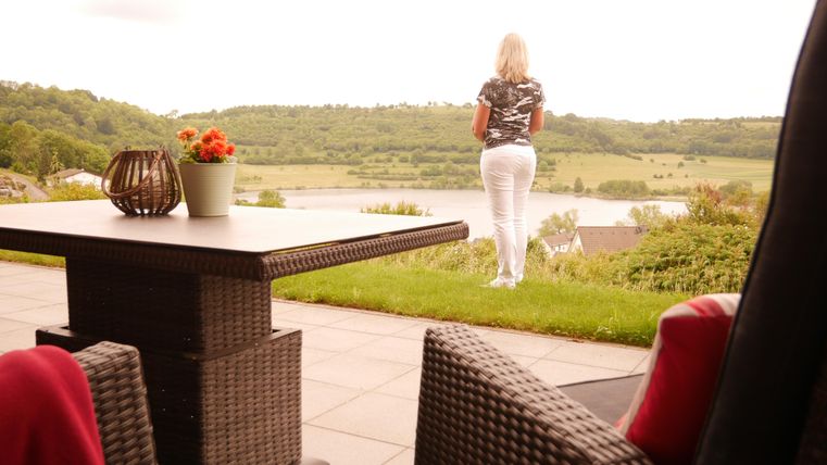Een vrouw staat op een terras en kijkt uit over een groene landschap met een waterpartij op de achtergrond. De tafel is versierd met een bloemenvaas en een decoratieve mand.