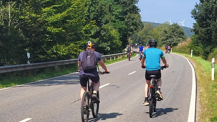 Twee fietsers rijden langs een rustige landweg. Op de achtergrond zijn er meer fietsers en groene bomen te zien.