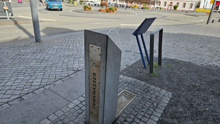 Une fontaine à eau en pierre se trouve sur une place pavée. En arrière-plan, des voitures et un panneau sont visibles.