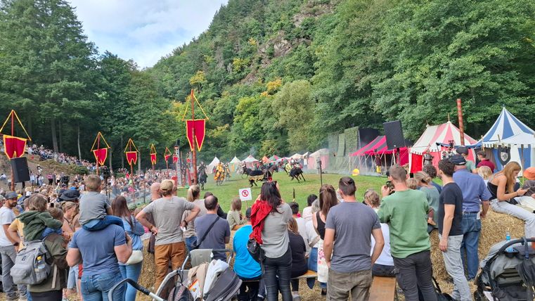 A large crowd is gathered on a meadow to watch an outdoor event. In the background, historical tents and a wooded landscape can be seen.