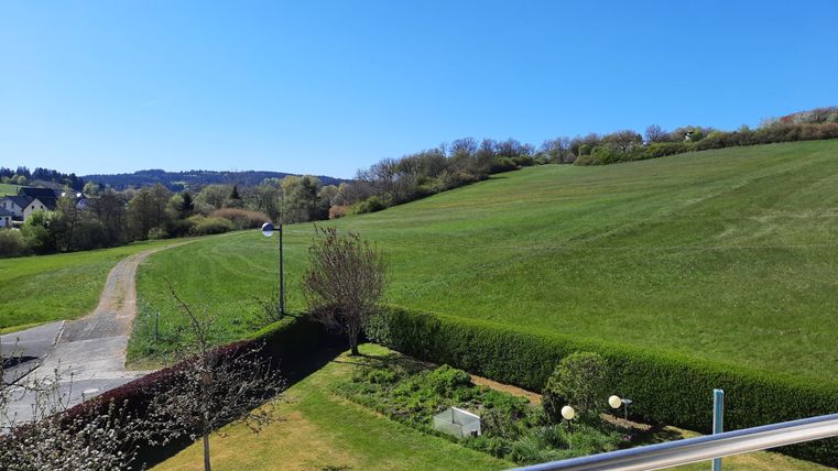 Eine grüne Landschaft mit sanften Hügeln und klarem blauen Himmel. Im Vordergrund sind Bäume und eine gepflegte Gartenfläche zu sehen.