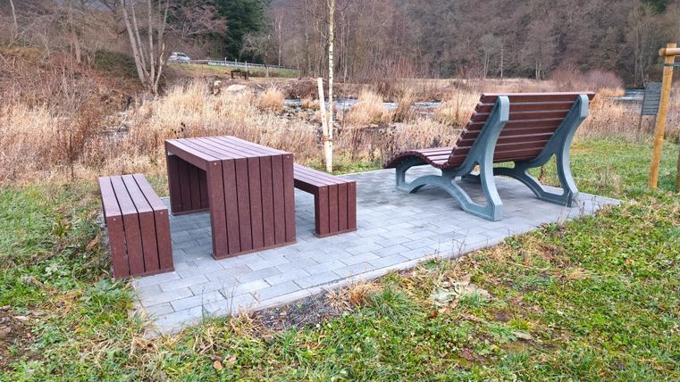 Eine Holzbank und ein Tisch stehen auf einem gepflasterten Bereich in der Natur. Im Hintergrund sind Bäume und Hügel zu sehen.